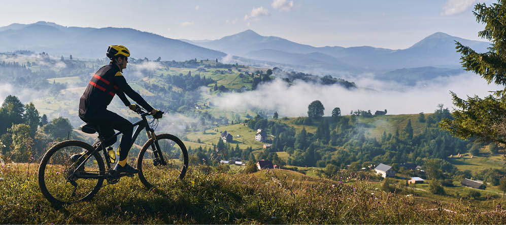 Man riding bicycle on grassy hill and looking at beautiful misty mountains. Male bicyclist enjoying panoramic view of majestic mountains during bicycle ride. Concept of sport, bicycling and nature.