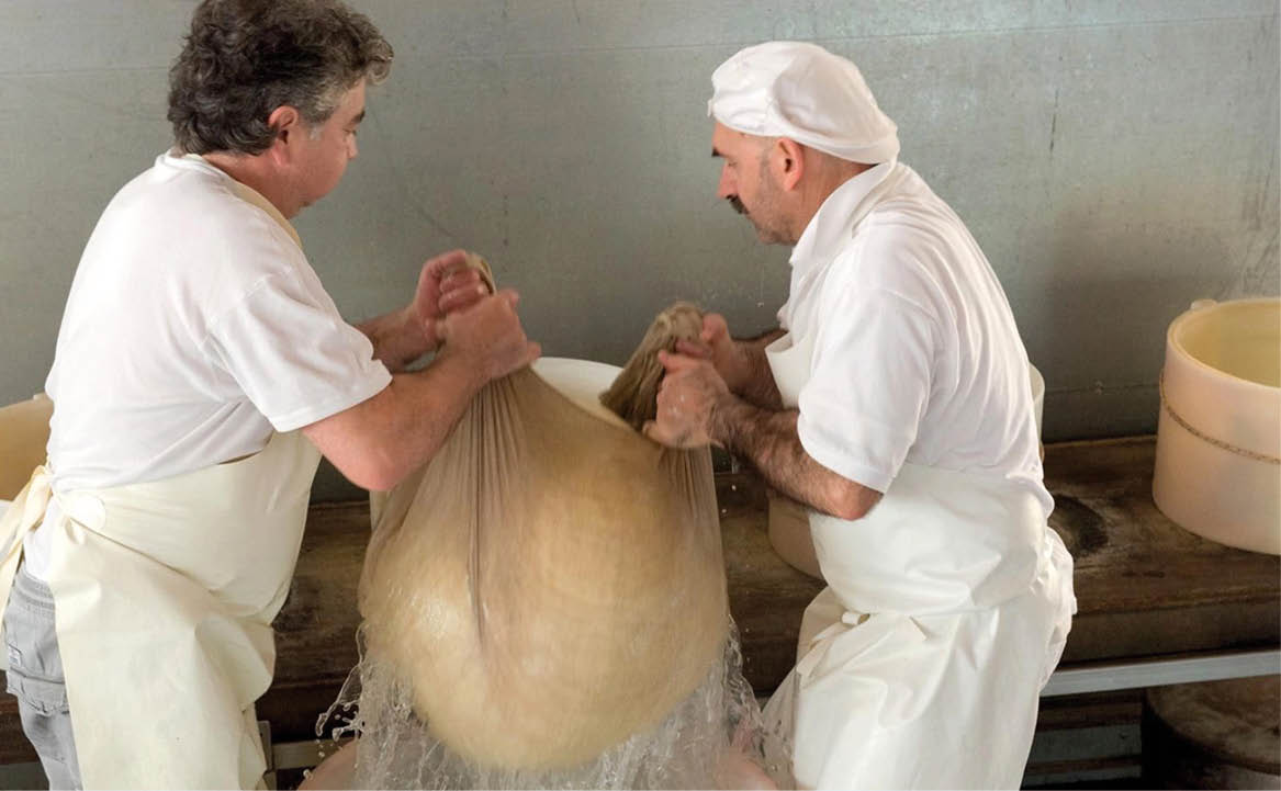 Zwei K ser heben gemeinsam geronnene K semasse in einem Tuch aus einem Kessel mit Molke.