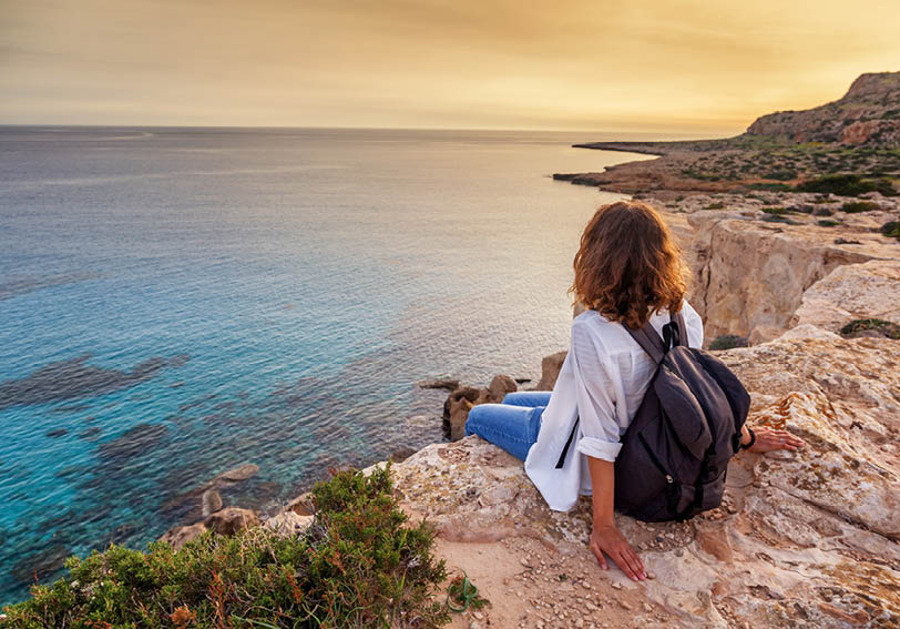A stylish young woman traveler watches a beautiful sunset on the rocks on the beach, Cyprus, Cape Greco, a popular destination for summer travel in Europe