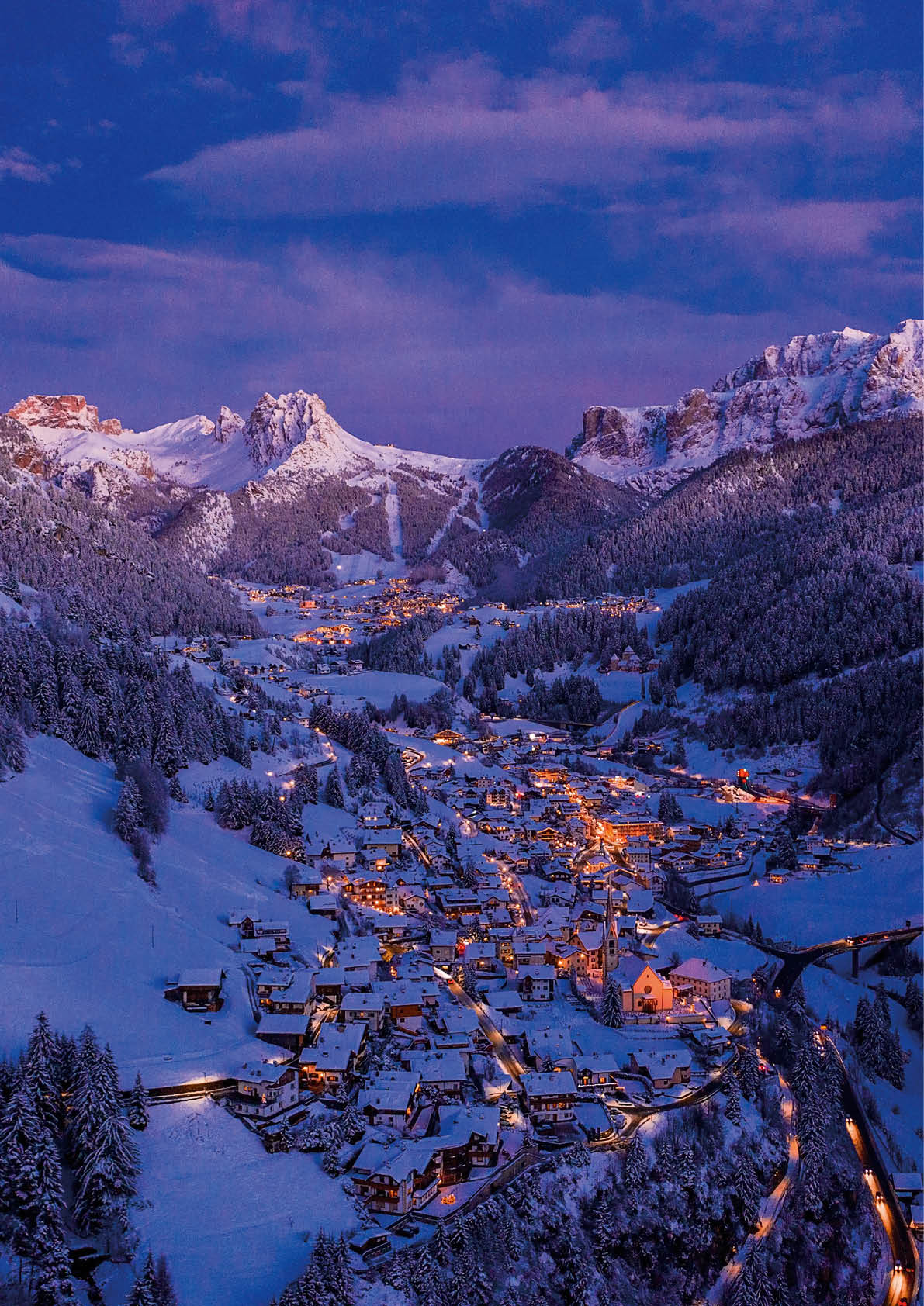 An aerial shot of a small bright town between snowy mountains during the evening