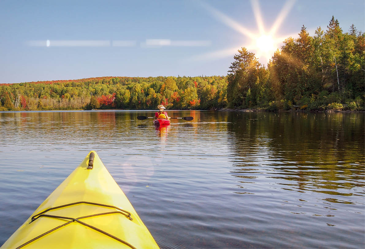 Perspektive aus einem gelben Kajak auf einen See mit herbstlich gef rbtem Wald und tief stehender Sonne.