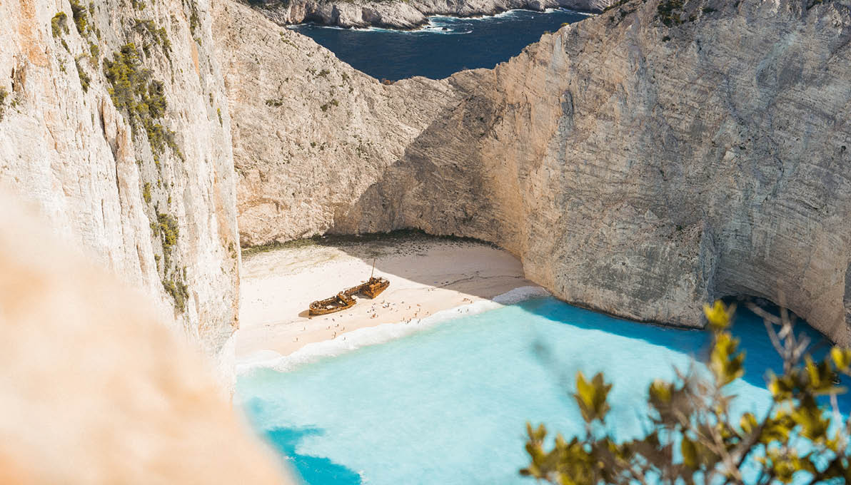 Famous shipwreck on Navagio beach with turquoise blue sea water surrounded by huge white cliffs. Famous landmark location on Zakynthos island, Greece. Farme shot between leaves.