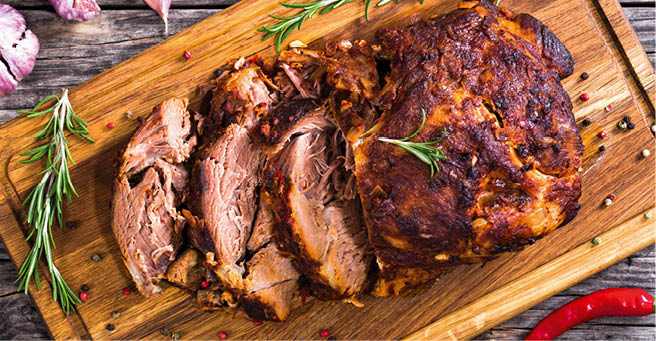 Oven-Barbecued Pork shoulder, cut on slices on chopping board with rosemary, peppercorn and bay leaf on old dark planks with garlic and chili on background, view from above, close-up