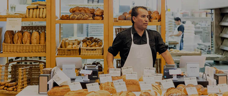 B ckereitheke mit verschiedenen Broten und Preisschildern, Verk ufer in Sch rze dahinter, Regale mit Backwaren im Hintergrund