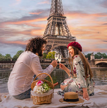 PARIS, FRANCE - Jun 07, 2019: A happy smiling couple toasts their love with wine and flowers on a picnic set. In front of them the Eiffel tower and the Seine river at sunset
