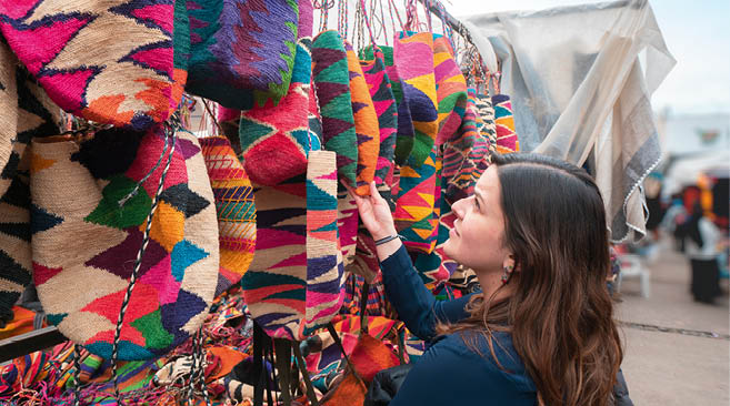 Beautiful young Latin American woman comparing Shigras that hang for sale in the Plaza de los Ponchos in the city of Otavalo. Shigra is a hand-woven indigenous bag