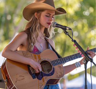 A woman in a cowboy hat is singing into a microphone while playing a guitar. She is surrounded by microphones and stands in front of a tree. Generated by AI