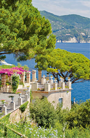 Villa mit bl hender Terrasse und Pinien ber dem Meer, wei e Yacht im Wasser, gr ne H gel im Hintergrund.