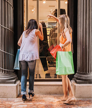 Zwei Frauen mit Einkaufstaschen betrachten ein Schaufenster mit Kleidung.
