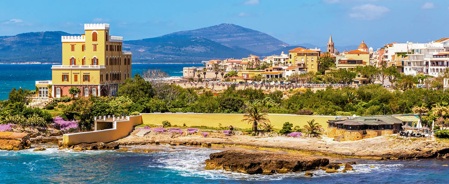 Mediterranean seacoast in Alghero city, Sardinia, Italy. Spring flowers and trees on foreground, colourful buildings of Alghero old city center on background