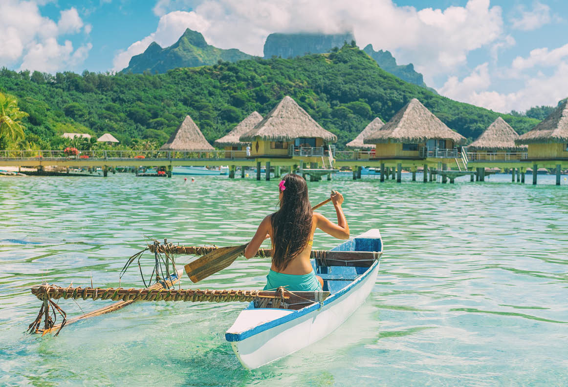 Bora Bora travel vacation iconic photo. Outrigger Canoe - woman paddling in traditional French Polynesian Outrigger Canoe. Mount Otemanu and overwater bungalow resort hotel sport lifestyle.