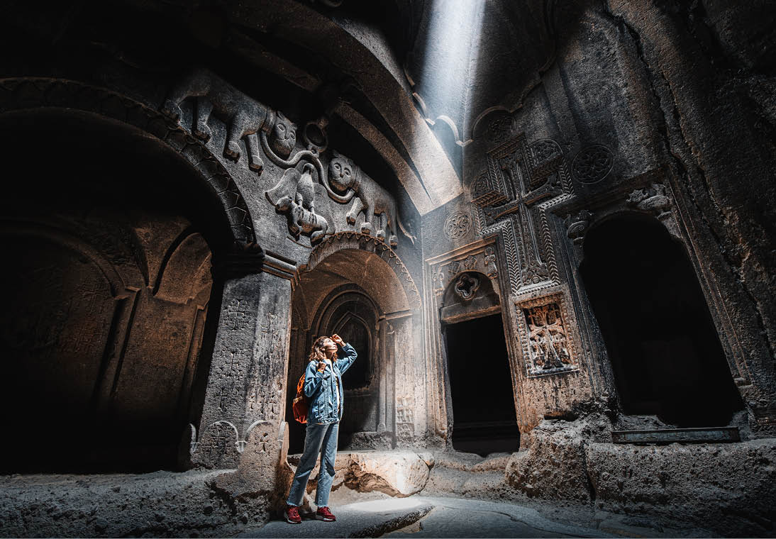 Woman tourist explores the mystical interior of the main hall of the Armenian Geghard Monastery. A ray of light falls on ancient bas-reliefs carved on the walls