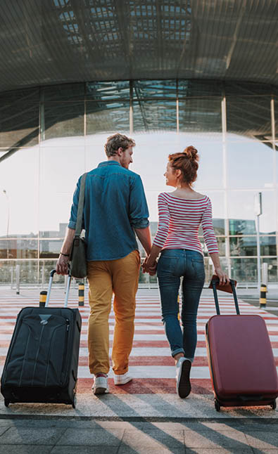 Full length back view portrait of young man and his charming girlfriend walking and carrying their trolley bags. They looking at each other and smiling