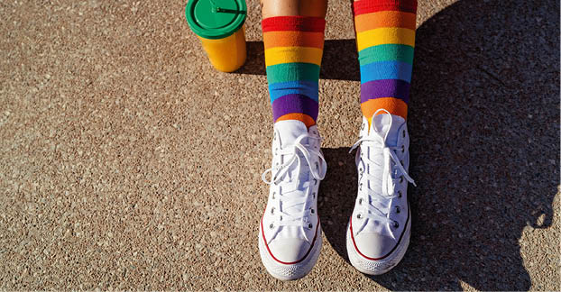 Top view of legs in bright colorful rainbow pride socks at sunlight outdoors. Trendy teen sitting on ground, generation Z lifestyle, pride month. 