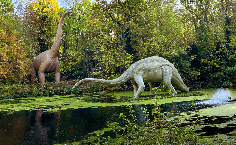 Zwei gro e Dinosaurierfiguren stehen am Wasser vor dichtem Laubwald.