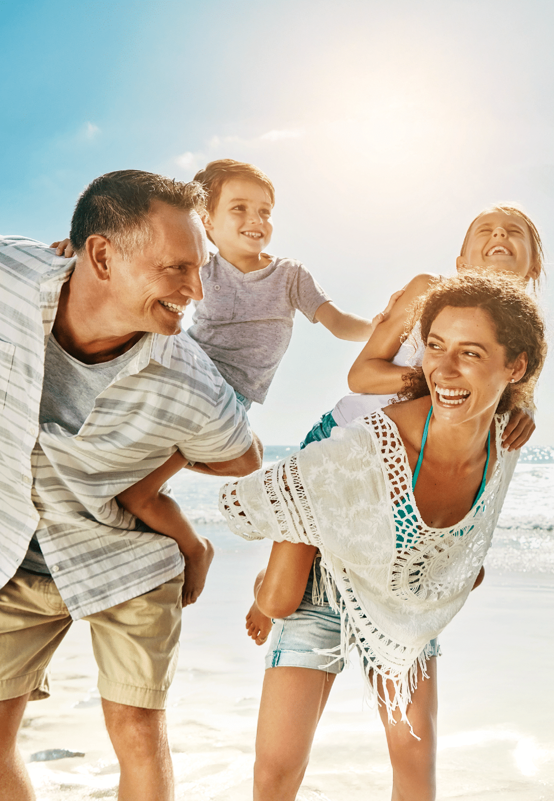 Lachende Familie spielt am Strand, Eltern tragen Kinder huckepack vor sonnigem Himmel und Meer.