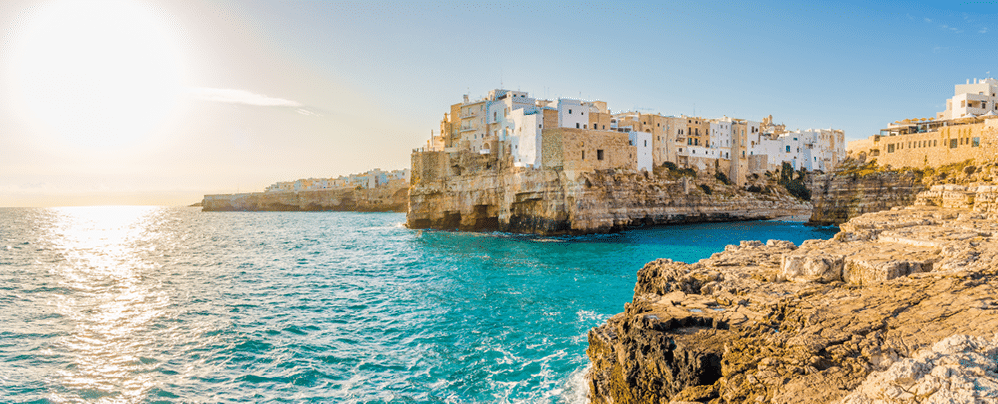 Sonnenaufgang in der N he der K ste von Polignano a Mare in Italien: Helle H user auf Felsklippe an t rkisfarbenem Meer, tiefstehende Sonne spiegelt sich im Wasser.