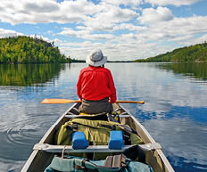 Person mit Paddel sitzt im beladenen Kanu auf einem ruhigen See zwischen bewaldeten Ufern.