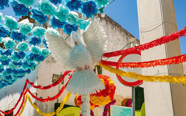 Papierdeko in Blau- und Rott nen sowie gro e wei e Taube h ngen ber schmaler Gasse bei Sonne.