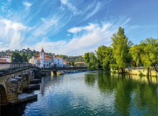 Tomar, Portugal: Steinbr cke mit Rundb gen, Kirche und Wasserfall in gr ner Flusslandschaft unter blauem Himmel.