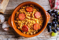 Tontopf mit Migas und Chorizo, daneben Knoblauch, Trauben, Brot und rot-wei es Tuch auf Holztisch.