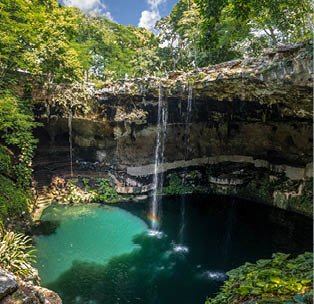 Cenote mit Wasserfall in Tropenlandschaft: Rundes Naturbecken mit Wasserfall, umgeben von Felsen und dichter Vegetation.