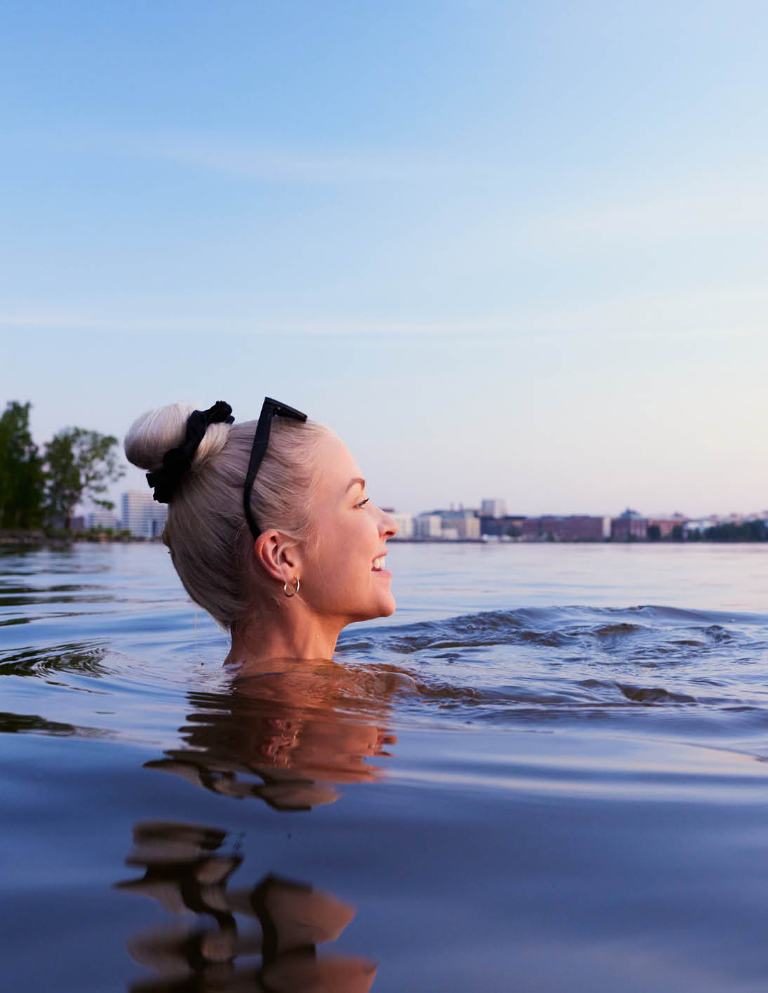 Blonde Frau mit Sonnenbrille im Wasser, Stadt im Hintergrund, Himmel in sanften Farbt nen.