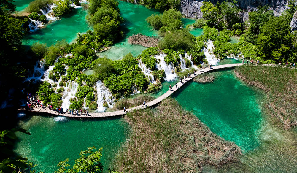 Holzsteg mit vielen Menschen zwischen Wasserf llen und klaren Seen in gr ner Umgebung.