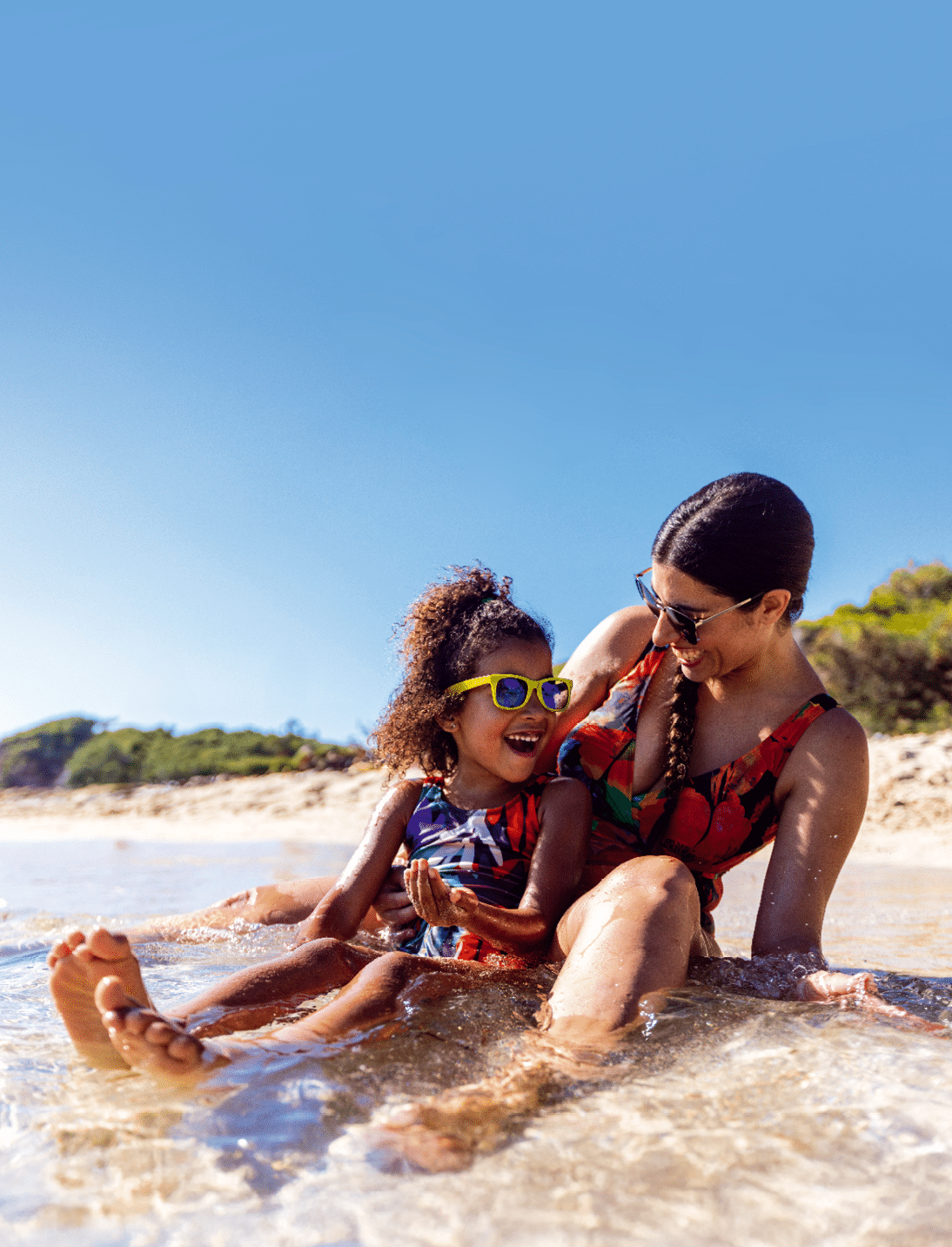 Frau mit Kind sitzt im flachen Wasser am Strand, beide tragen Sonnenbrillen und lachen.