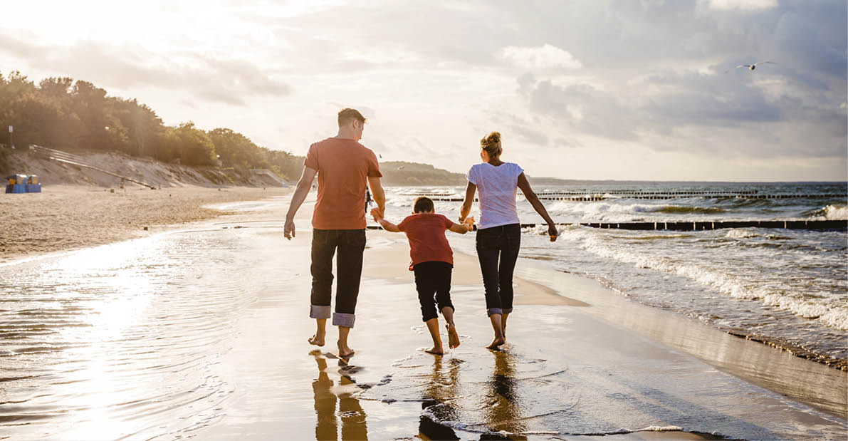 Zwei Erwachsene und ein Kind bei Sonnenuntergang auf nassem Sandstrand mit ruhiger Brandung.