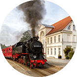 B derbahn Molli in Bad Doberan: Schwarze Dampflok mit roten Waggons st t Rauch aus und steht vor wei em Geb ude mit rotem Dach.