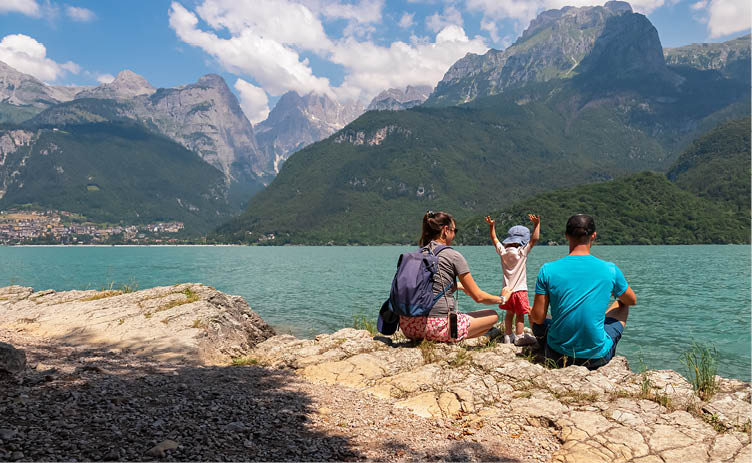 Junge Familie mit kleinem Kind sitzen im Sommer am Ufer des Alpensees Lago di Molveno, umgeben von Felsen und Berggipfeln der Brenta-Dolomiten.