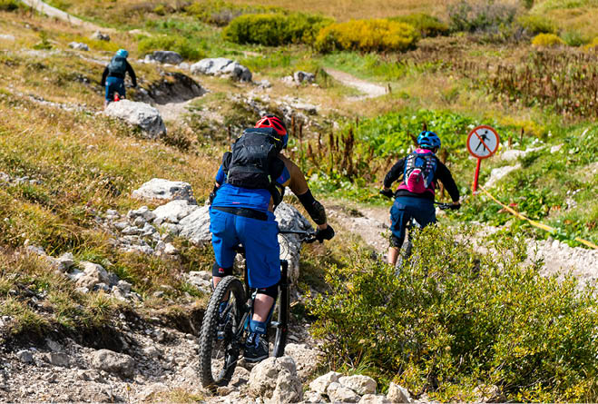Drei Personen mit Helmen fahren mit Mountainbikes auf schmalem Pfad durch offene Landschaft.