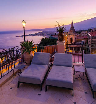 Gepolsterte Liegen auf Terrasse mit Blick auf Stadt, Meer und Berge bei Sonnenuntergang.