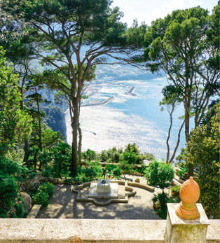 Steinmauer mit Brunnenanlage und dichtem Baumbestand ber einer glitzernden Bucht.