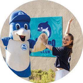 Frau klatscht mit einem Delfin-Maskottchen in blauer Kleidung vor einem Schild mit Flosse-Club-Logo ab.