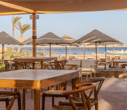 Holztische und Sonnenschirme auf gepflasterter Terrasse mit Blick auf Sandstrand und Steg.