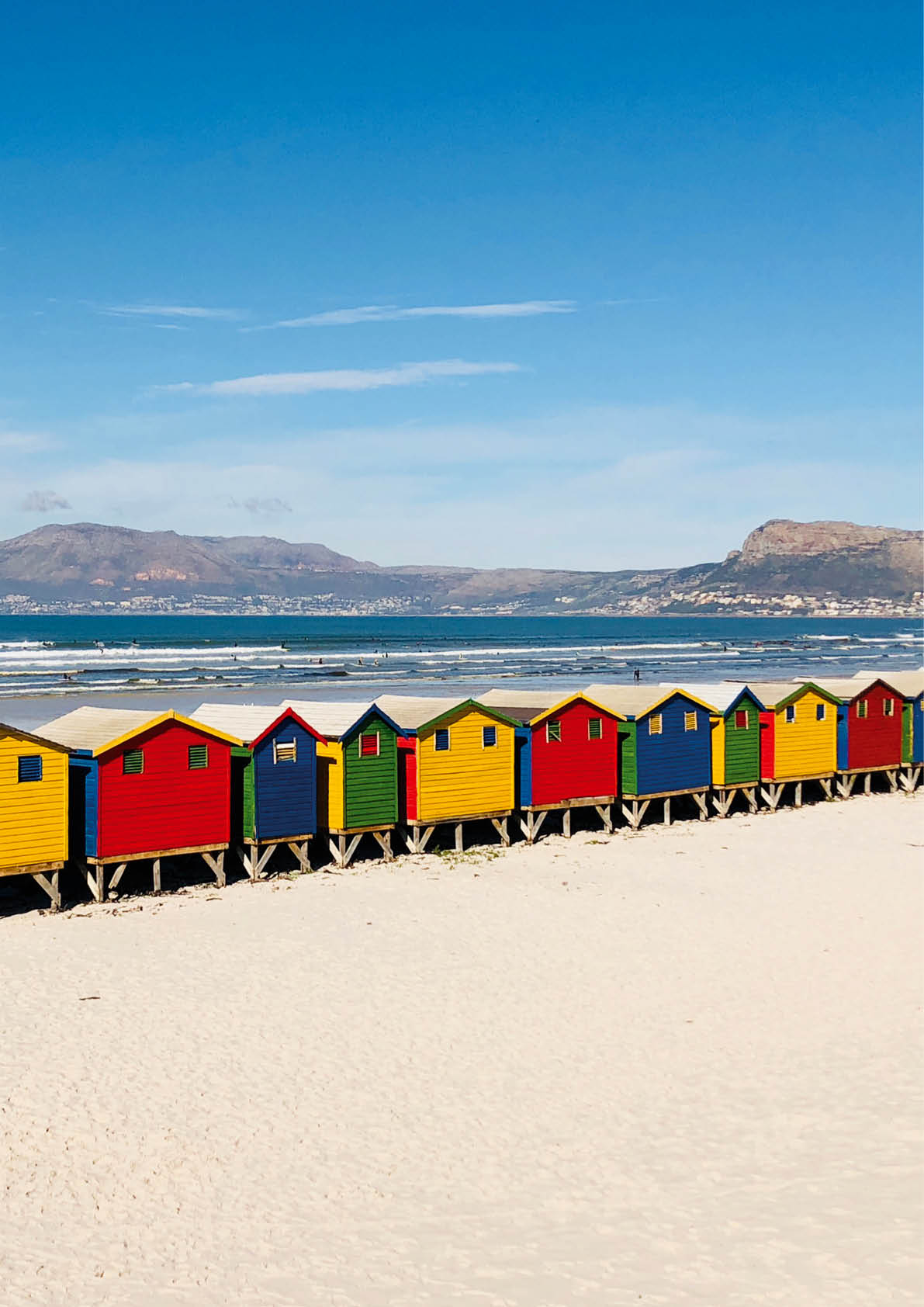 Reihe bunter Holzh tten auf hellem Sandstrand mit Blick auf Meer und Berge im Hintergrund.