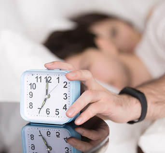 Young couple awaking after wake-up signal in the morning at home. Sleepy man stretching hand to alarm clock touching button to snooze or turn it off. Focus on hand with clock. Time to wake up