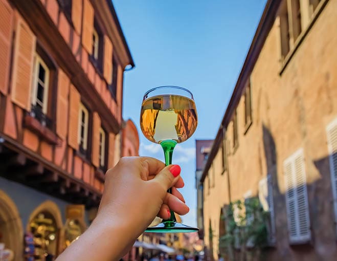 Woman s hand with a glass of Gewurztraminer wine at an outdoor restaurant, blurred half timbered houses in Colmar France, Alsace Wine Route village