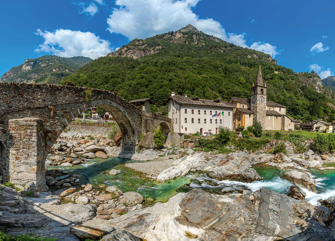 Panorama of an old stone bridge over an alpine river with rocks, as a small church stands against a mountain in the background in Lillianes, Aosta Valley, Italy.