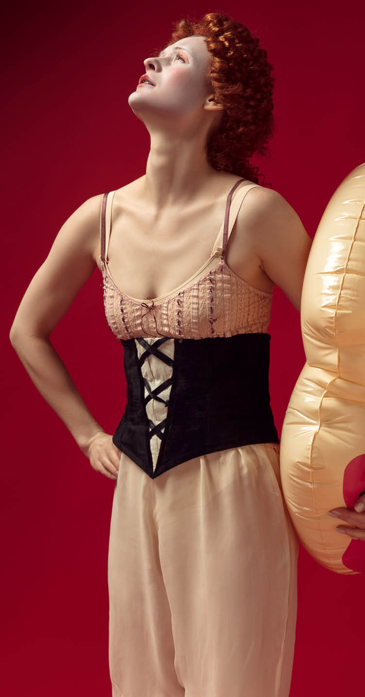 Medieval redhead young woman as a duchess in black corset and night clothes standing on red background with a swim circle as a donut. Concept of comparison of eras, modernity and renaissance.