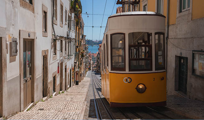 A classic yellow tram navigates a steep, narrow street in Lisbon, Portugal, with traditional European buildings and a glimpse of the Tagus River in the background.