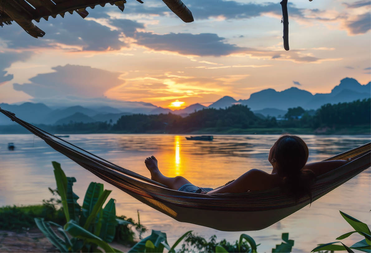 Person liegt in H ngematte am Flussufer und blickt auf Sonnenuntergang ber dem Mekong mit Silhouetten von Bergen im Hintergrund. 