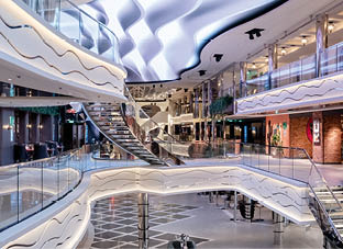 Modernes, mehrst ckiges Innenraumdesign auf einem Kreuzfahrtschiff mit geschwungenen Gel ndern, wellenf rmiger Beleuchtung an der Decke, offenen Sitzbereichen und Gesch ften im Hintergrund. 