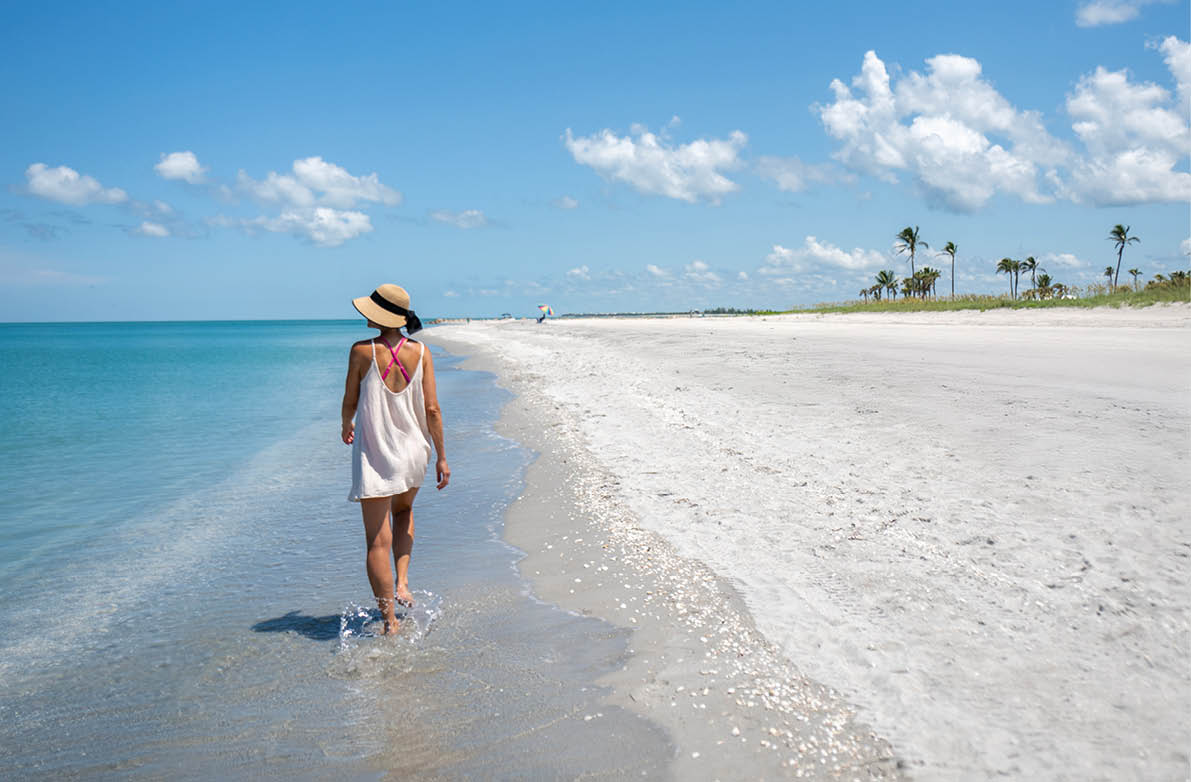 Person geht am Rand eines leeren Sandstrandes im seichten Wasser entlang, im Hintergrund Palmen und blauer Himmel.