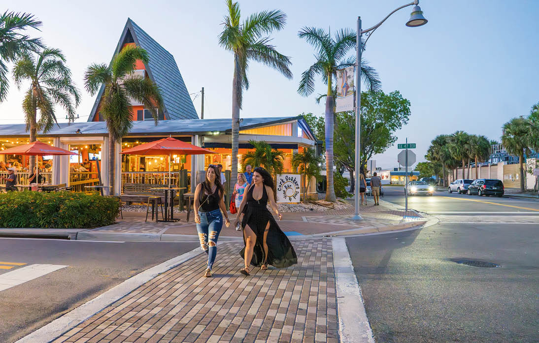 Zwei Frauen berqueren eine Stra e vor einem Restaurant mit Au enterrasse, Palmen und Abendbeleuchtung
