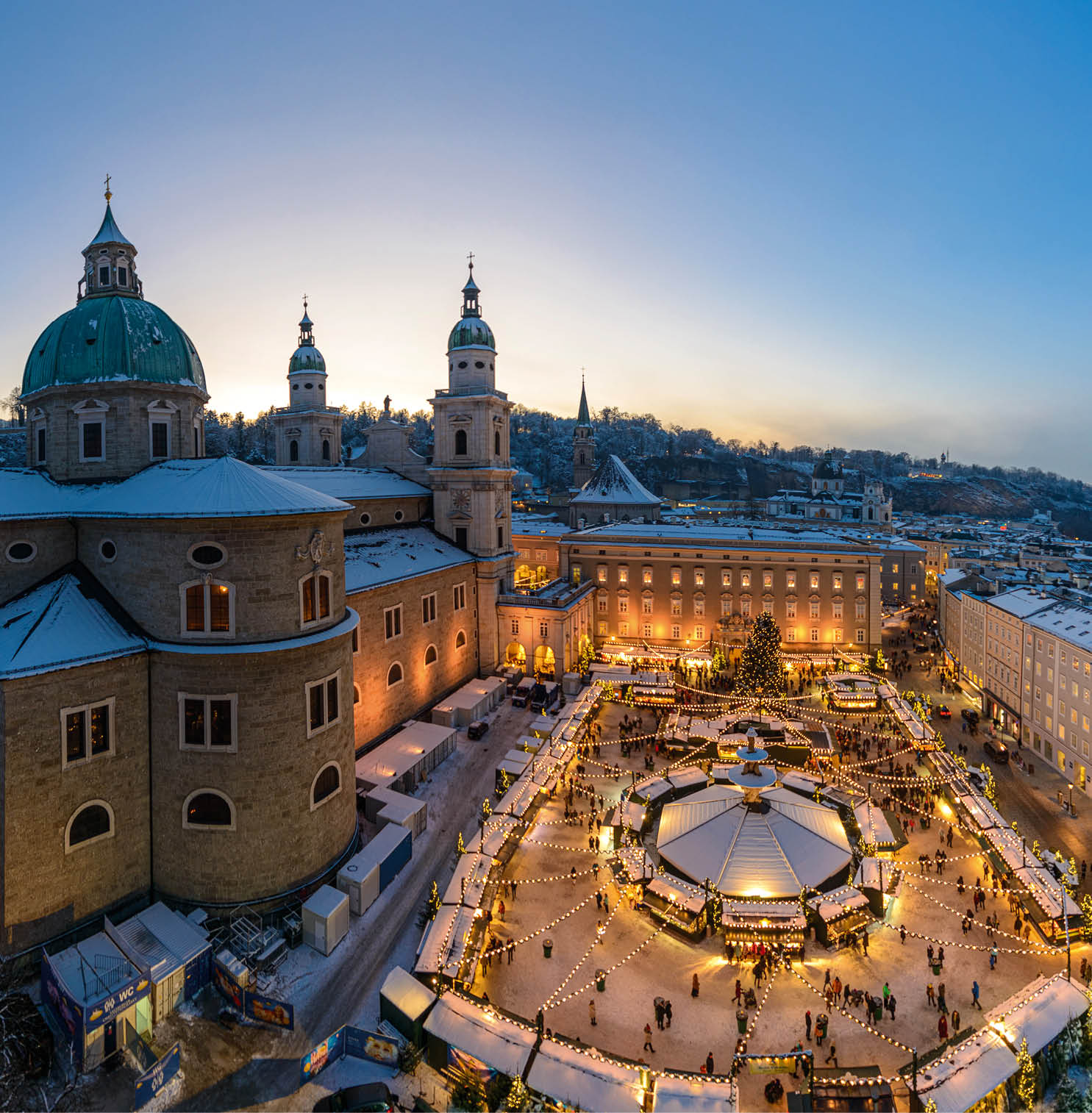 Beleuchteter Weihnachtsmarkt auf dem verschneiten Domplatz in Salzburg mit Festung Hohensalzburg im Hintergrund.