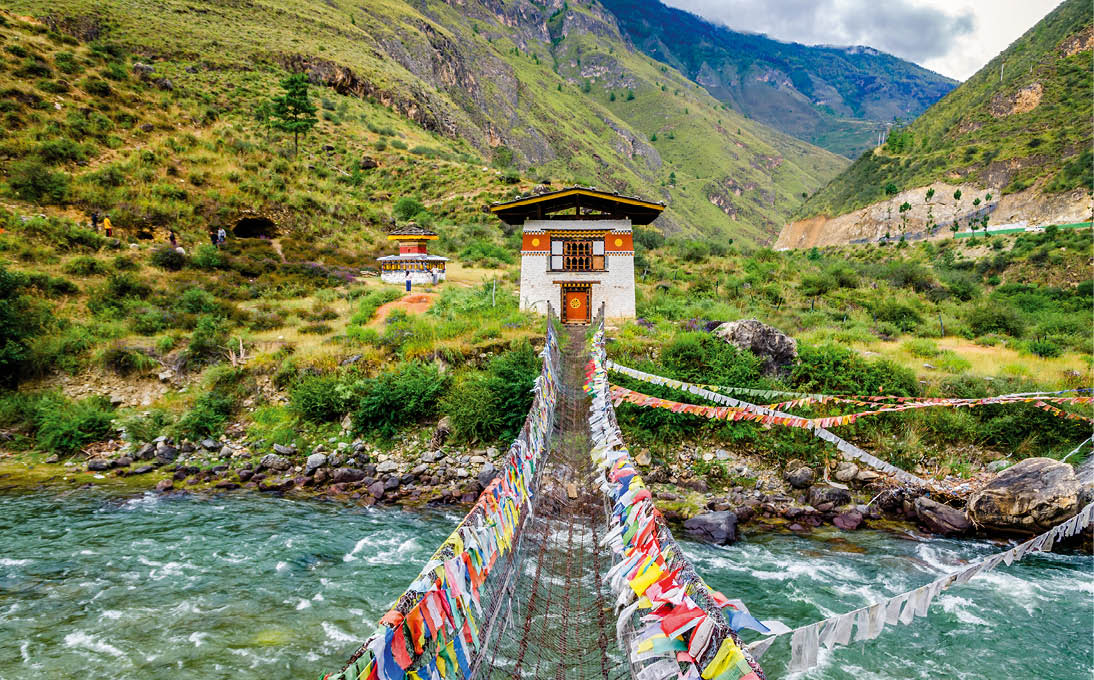 H ngebr cke mit bunten Gebetsfahnen f hrt ber Fluss zu traditionellem Geb ude in gr nem Bergtal.