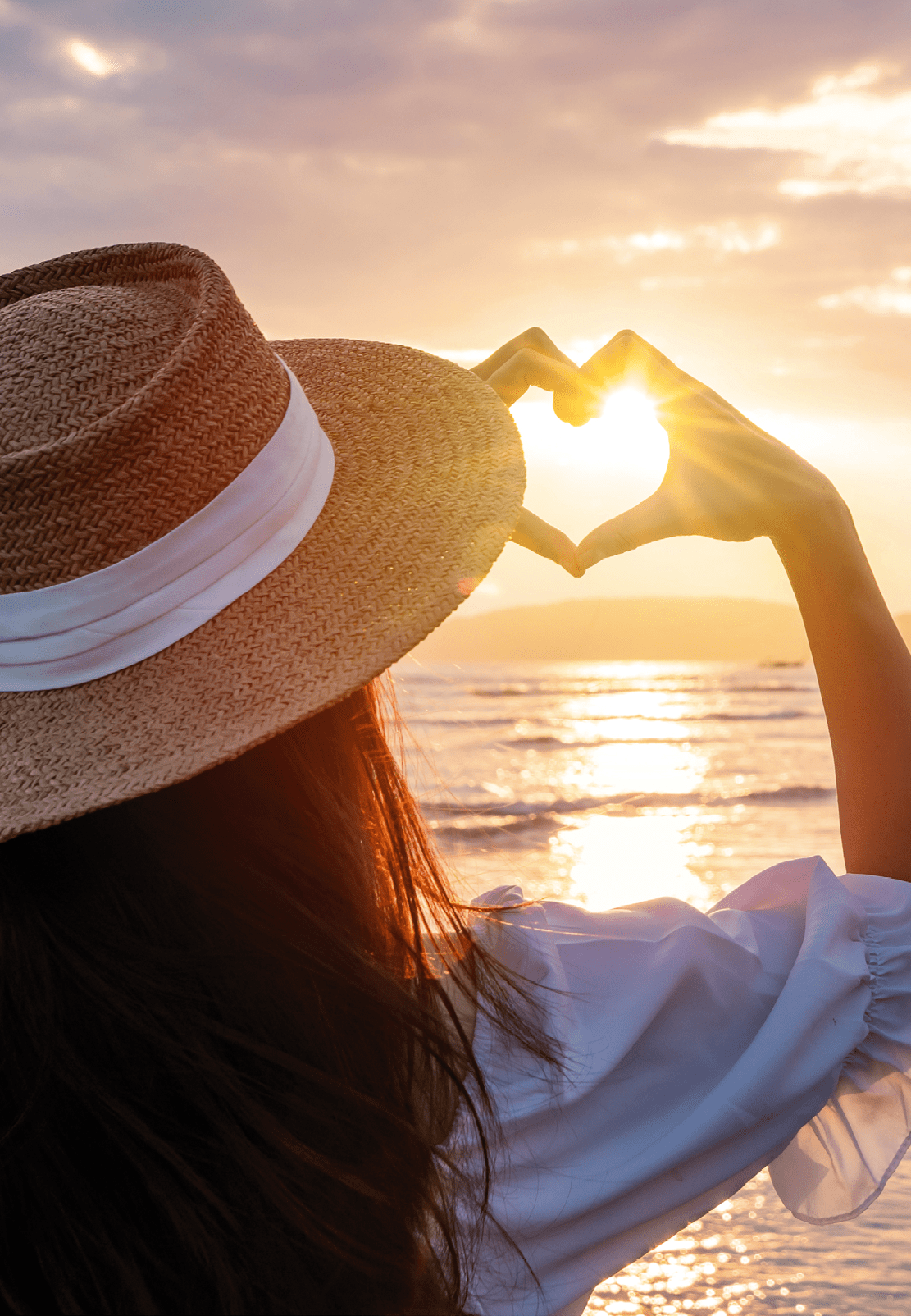 Frau mit Strohhut formt mit den H nden ein Herz vor der untergehenden Sonne am Meer.